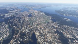 An aerial view shows dense urban neighborhoods, industrial buildings, and curving highways surrounded by forests and expansive lakes dotted with islands.