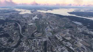 An aerial view shows dense neighborhoods, highway interchanges, and industrial buildings beside wooded lakes and islands beneath pink clouds.