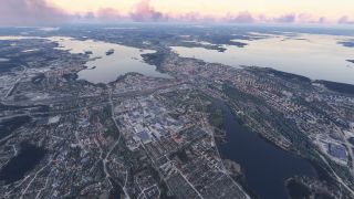 A city spreads between broad lakes, with dense neighborhoods, industrial buildings, and wooded islands beneath a pink-tinged cloud bank.