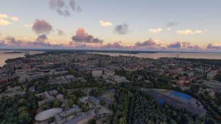 A waterfront city spreads beneath pink clouds, with dense buildings, tree-lined parks, and blue outdoor pools visible from above.