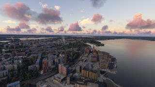 A waterfront city extends beside a broad lake, with apartment blocks, smoking chimneys, and a distant observation tower beneath pink clouds.