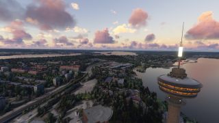 An illuminated observation tower overlooks a lakeside city with tree-lined roads and low-rise buildings beneath pink and orange clouds.