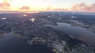 An aerial view shows dense urban neighborhoods between large lakes, with wooded shorelines, connecting roads, and pink clouds along a golden horizon.