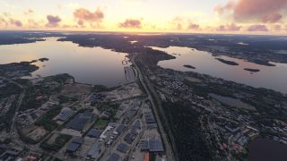 An aerial view shows industrial buildings, residential neighborhoods, and roads between broad waterways dotted with wooded islands beneath an orange, cloud-filled sky.