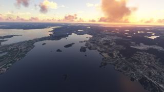 An aerial view shows broad waterways dotted with wooded islands, shoreline neighborhoods, and winding roads beneath orange clouds and a low sun.