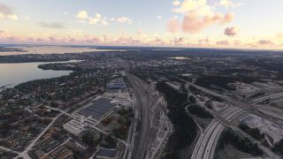 An aerial view shows a lakeside city with extensive railway tracks, highway interchanges, warehouses, and wooded neighborhoods beneath pink clouds.