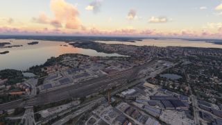 An aerial view shows an extensive railway yard, industrial buildings, and lakeside neighborhoods beneath scattered pink and gold clouds.