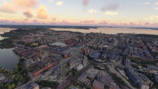 An aerial view shows a lakeside city with an oval arena, railway tracks, and dense urban blocks beneath pink clouds.