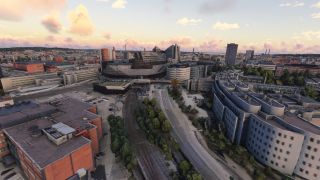Nokia Arena stands among city buildings, with railway tracks and a tree-lined road running through the foreground beneath pink clouds.