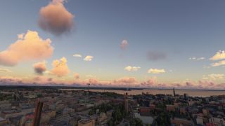 A waterfront city spreads beneath pink and orange clouds, with industrial chimneys, dense buildings, and a slender observation tower.