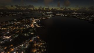 Illuminated streets and waterfront buildings surround dark waterways at twilight, with a blue-lit tower rising beneath scattered clouds.