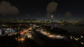 A blue-lit observation tower rises above a waterfront city, with illuminated roads, shoreline buildings, and scattered clouds in the dark sky.