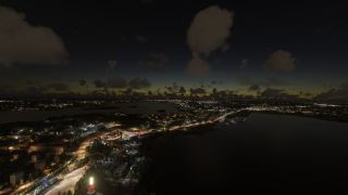 Illuminated roads and buildings line dark waterways beneath scattered clouds and a faint orange horizon in an aerial twilight view.