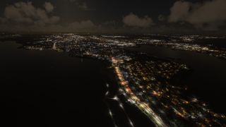 An aerial nighttime view shows illuminated waterfront roads and dense city lights bordering dark waterways beneath scattered clouds.