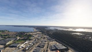 An aerial view shows industrial buildings and parallel roads beside wooded neighborhoods, with broad waterways and small islands stretching toward the horizon.
