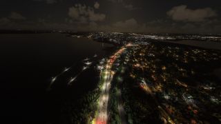 An aerial nighttime view shows illuminated shoreline roads, marina piers, and residential neighborhoods beside dark water under scattered clouds.