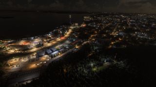 An aerial nighttime view shows illuminated roads, parking lots, and buildings beside dark water, with a blue-lit tower near the shoreline.