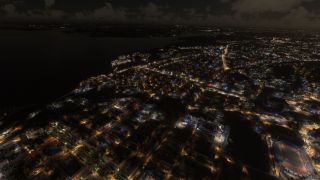 A nighttime aerial view shows illuminated city streets beside dark water, with a stadium in the foreground and clouds overhead.