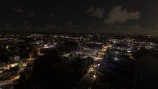 An aerial nighttime view shows illuminated city streets, a floodlit sports stadium, and bridges crossing dark waterways beneath scattered clouds.