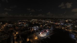 A nighttime aerial view shows illuminated waterfront buildings, docked boats, and an industrial chimney beneath scattered clouds.
