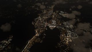 An aerial nighttime view shows networks of white and amber city lights interrupted by dark areas and scattered clouds.