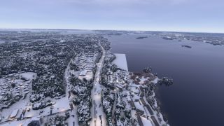 Snow-covered neighborhoods and illuminated roads line a broad lake, with wooded islands scattered across the water beneath a pale sky.