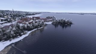 Snow-covered trees and red-roofed buildings line a curved shoreline beside dark water, with wooded islands visible in the distance.