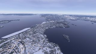 Snow-covered neighborhoods and winding roads spread across a peninsula surrounded by dark water and small wooded islands beneath an overcast sky.