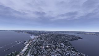 A snow-covered town stretches along a peninsula, with illuminated roads, waterfront docks, and broad waterways beneath an overcast sky.