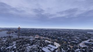An illuminated observation tower rises above a snow-covered city beside a lake, with roads winding between buildings beneath layered clouds.