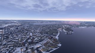A snow-covered city borders broad waterways, with illuminated waterfront buildings, industrial chimneys, and a tall tower beneath a clouded sky.