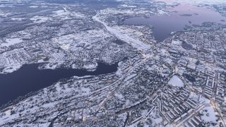 Snow covers dense residential neighborhoods, industrial buildings, and forests surrounding dark waterways, while illuminated roads wind through the town.