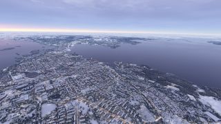 A snow-covered city stretches along broad waterways, with illuminated streets, wooded shorelines, and distant islands beneath a cloudy sky.