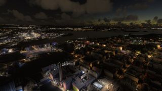 An aerial view shows illuminated city blocks, waterfront industrial chimneys, and an oval stadium beneath a dark, cloud-filled sky.
