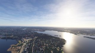 An aerial view shows lakeside neighborhoods, curving roads, and wooded islands, with bright sunlight reflecting across the water.