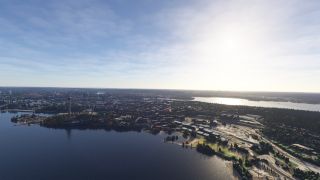A lakeside city stretches between broad waterways, with a tall observation tower, wooded shorelines, industrial buildings, and curving roads.