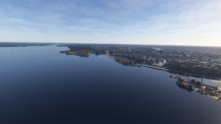 A waterfront city stretches along a broad blue lake, with shoreline roads, industrial chimneys, and wooded peninsulas visible from above.
