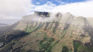 Low clouds cross a rocky mountain ridge above green slopes and winding trails, with coastal buildings and ocean visible to the left.