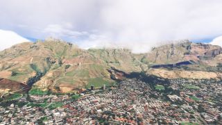 Dense neighborhoods and winding roads spread beneath a rocky mountain ridge, with low clouds obscuring sections of its flat summit.