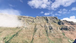 A broad, flat-topped mountain ridge rises above rocky slopes and winding trails, with low cloud obscuring its left flank.