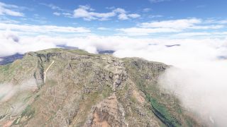 A flat-topped mountain rises above steep, layered rock cliffs and green slopes, with low clouds partially obscuring the surrounding terrain.