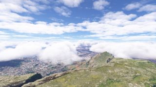 A rocky, grass-covered mountain ridge overlooks dense urban streets, while low white clouds partially obscure the city beneath a blue sky.