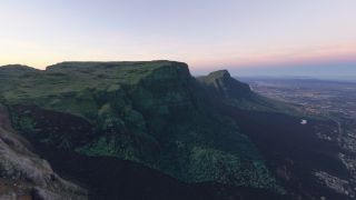 A flat-topped mountain rises above forested slopes, with steep rocky cliffs overlooking an expansive city beneath a pink and blue sky.