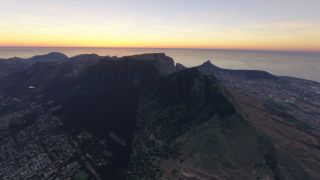 Table Mountain rises above dense urban neighborhoods, with winding hillside trails and an ocean horizon beneath a yellow-orange sky.