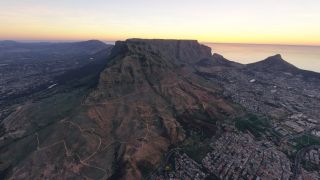 Table Mountain rises above dense urban neighborhoods, with winding trails across its slopes and the ocean beneath a yellow-orange sky.