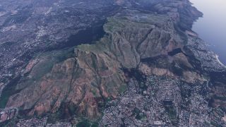 An aerial view shows steep, flat-topped mountains surrounded by dense urban neighborhoods, with winding hillside trails and a coastline to the right.
