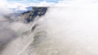 Low clouds partially obscure a flat-topped mountain with steep rocky cliffs, while coastal buildings and blue water appear beyond its slopes.