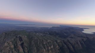 Steep, rocky mountain cliffs overlook a sprawling coastal city and curved bays beneath a pink and yellow sky.