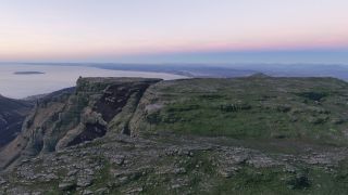 A rocky, flat-topped mountain overlooks a coastal city and offshore island beneath a pale pink sky.
