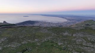 A rocky, shrub-covered plateau overlooks a coastal city, harbor, and broad bay with an offshore island beneath a pink sky.